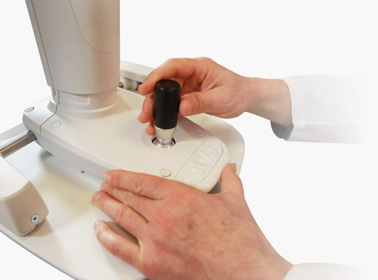 A Person In A White Lab Coat Is Skillfully Operating A Nidek Laboratory Device, Using Its Black Joystick-Like Handle And Multiple Buttons On The Base. This Advanced Equipment Seems To Be Designed For Precise Adjustments Or Measurements In A Scientific Or Medical Setting.