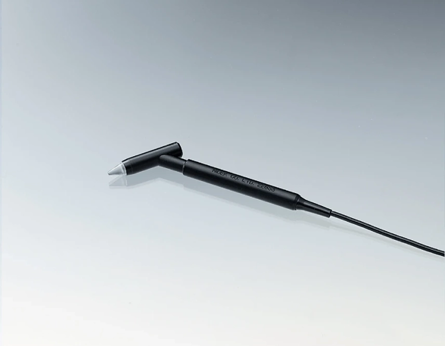A Black Handheld Medical Device By Nidek With A Long, Slender Body And A Small, Angled Tip, Placed Against A Neutral, Light-Colored Background. A Cord Extends From The End Of The Device Opposite The Angled Tip.