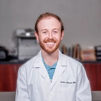 A Smiling Man With A Beard And Short, Red Hair Is Sitting And Wearing A White Lab Coat Over A Blue Checkered Shirt. The Nidek Lab Coat Has A Name Tag On The Left Side. He Is Seated Indoors, With A Blurred Background Featuring Office Equipment And Shelves Of Books.