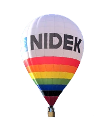 A Hot Air Balloon With &Quot;Nidek&Quot; Written On It In Large Black Letters Floats Gracefully. The Balloon Boasts Horizontal Stripes In Rainbow Colors—From Red And Orange To Yellow, Green, Blue, And Violet. Below, A Basket Filled With Passengers Enjoys The Serene Ride.