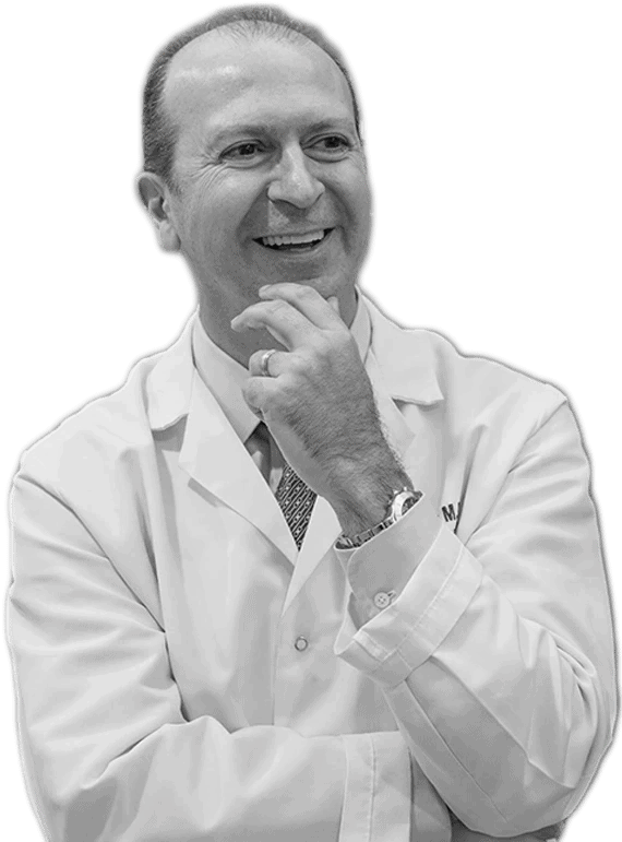 A Man In A White Medical Coat, Representing Nidek, Smiles And Gestures With His Hand Near His Chin. He Is Looking Off To The Side Against A Plain Gray Background.