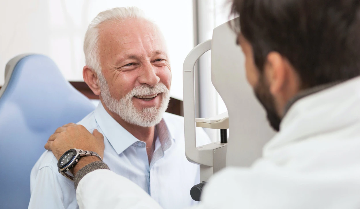 An Older Man With White Hair And A Beard Is Smiling While Sitting In An Eye Examination Chair. A Male Optometrist With Dark Hair Is Examining Him Using Nidek Eye Testing Equipment And Has His Hand Reassuringly On The Patient'S Shoulder.