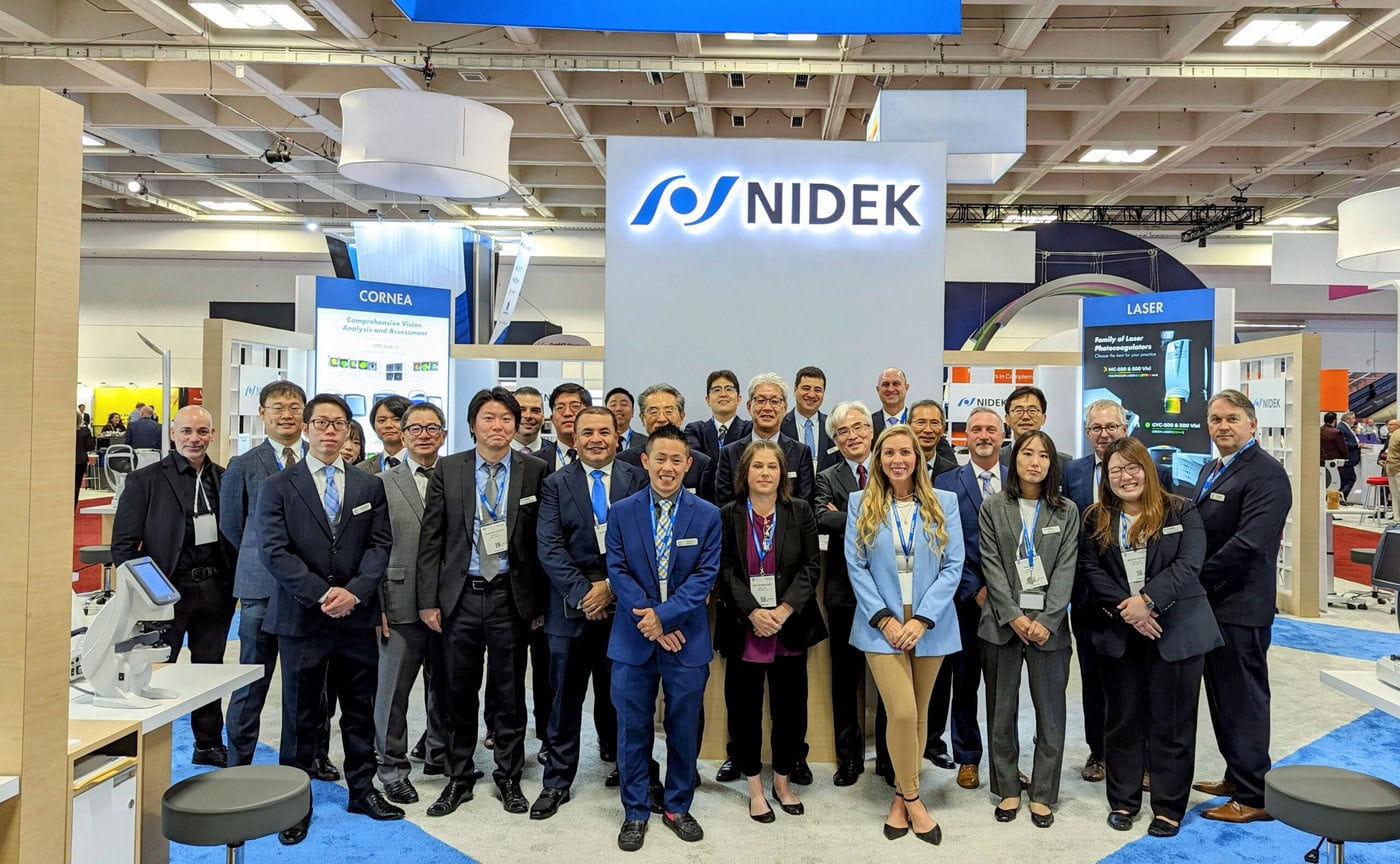 A Group Of People Stands In Front Of A Nidek Booth At An Exhibition. They Are Dressed In Business Attire, Smiling, And Facing The Camera. The Booth Features Various Displays Related To Nidek'S Medical Equipment And Technology. The Setting Is A Large, Well-Lit Convention Hall.