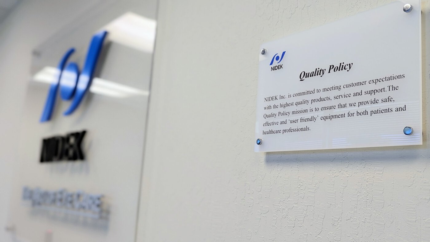 A Clear Plaque On A Wall Displays Nidek'S Quality Policy, Emphasizing The Commitment To Meeting Customer Expectations With High-Quality Products, Service, And Support. The Blurred Nidek Company Logo And Name Are Visible On The Adjacent Wall In The Background.