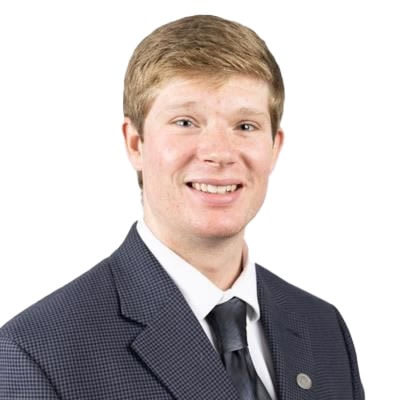 A Young Man With Short, Light Brown Hair Smiles At The Camera. He Is Wearing A Dark, Checkered Suit Jacket, A White Collared Shirt, And A Black Tie. The Background Is Plain White.