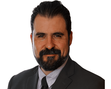 A Man With Dark Hair And A Beard Is Wearing A Dark Suit, White Shirt, And Striped Tie. He Is Smiling Slightly And Facing The Camera Against A Plain White Background.