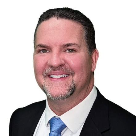 A Man With Short, Dark Hair, A Goatee, And A Mustache Is Smiling At The Camera. He Is Wearing A Black Suit Jacket, A White Dress Shirt, And A Light Blue Tie. The Background Is Plain White.