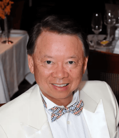 A Man Wearing A White Tuxedo Jacket And A Patterned Bow Tie Smiles At The Camera In A Formal Dining Setting With Tables, Glasses, And Plates In The Background.