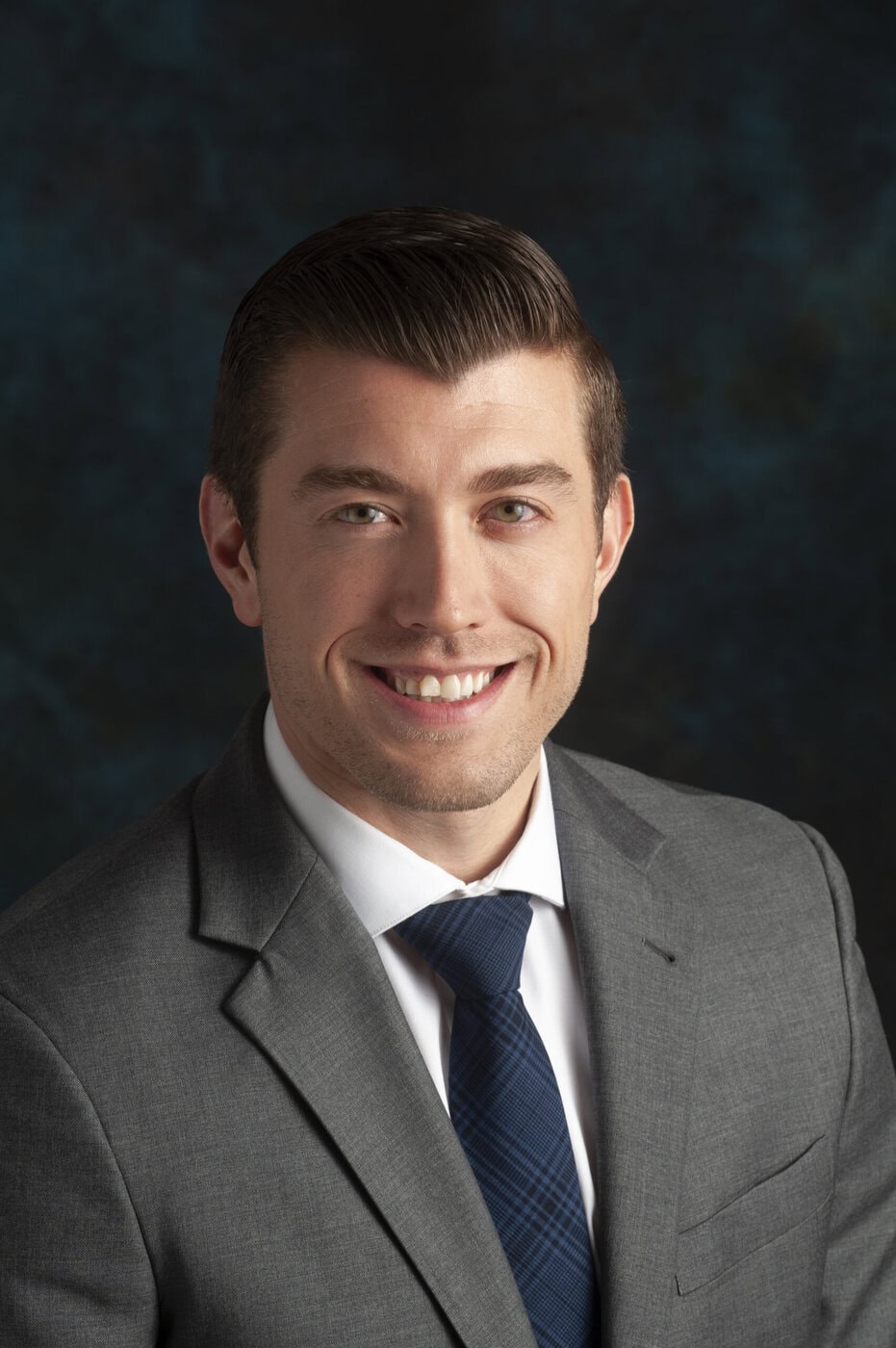 A Young Man With Short Brown Hair, Wearing A Gray Suit, White Shirt, And Blue Patterned Tie, Smiles At The Camera Against A Dark, Mottled Background.