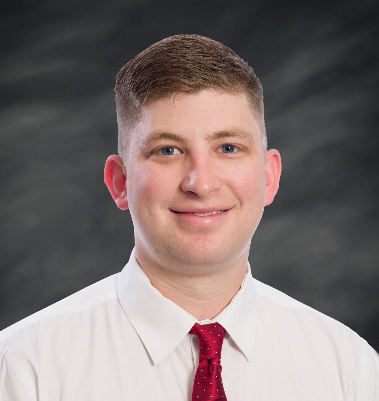 A Man With Short Light Brown Hair, Wearing A White Dress Shirt And A Red Tie With Small Dots, Is Smiling In Front Of A Dark, Blurred Background.
