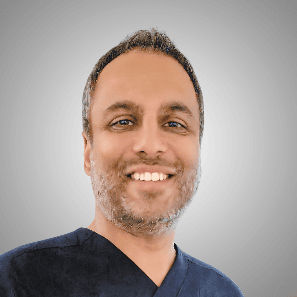 A Man With Short Hair And A Trimmed Beard Smiles At The Camera, Wearing A Dark V-Neck Shirt, Set Against A Light Gray Gradient Background.